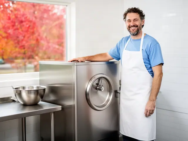 Smiling ice-cream shop owner in a blue T-shirt and white apron rests his hand on a stainless-steel batch freezer