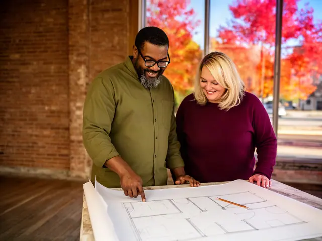 Two business partners in a brick-walled space smiling over a large floor plan spread on a table